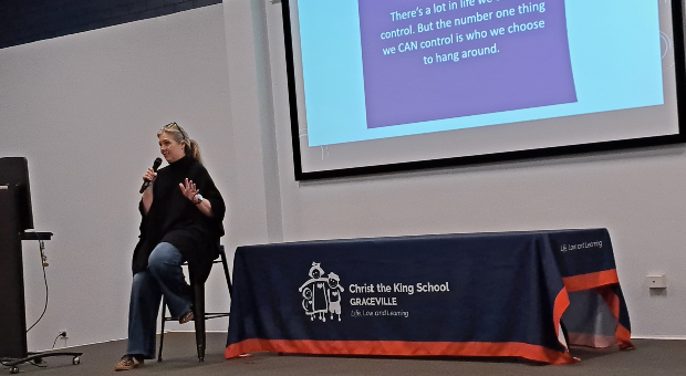 Rebecca Sparrow presents to Christ the King School families, seated on stage beside a presentation screen displaying friendship advice during the 6 Lessons in Friendship seminar.