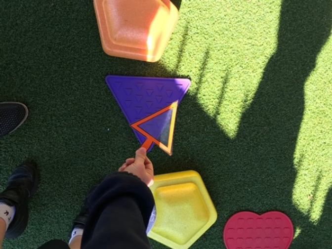 Prep student uses a triangular shape frame to match playground equipment, identifying 2D shapes during an outdoor mathematics investigation.