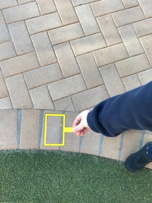 Prep student places a rectangular shape frame over pathway pavers, identifying and comparing geometric shapes around school grounds.